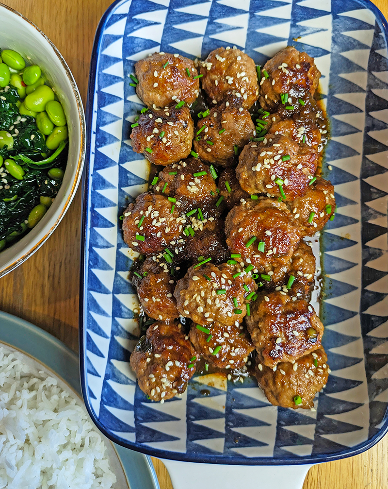 Sticky Chinese five spice meatballs in a blue and white dish. The meatballs look caramelised and glistening with the sauce. Decorated with sesame seeds and chives.